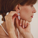 Woman wearing heart-shaped earrings and a pearl necklace against a neutral background