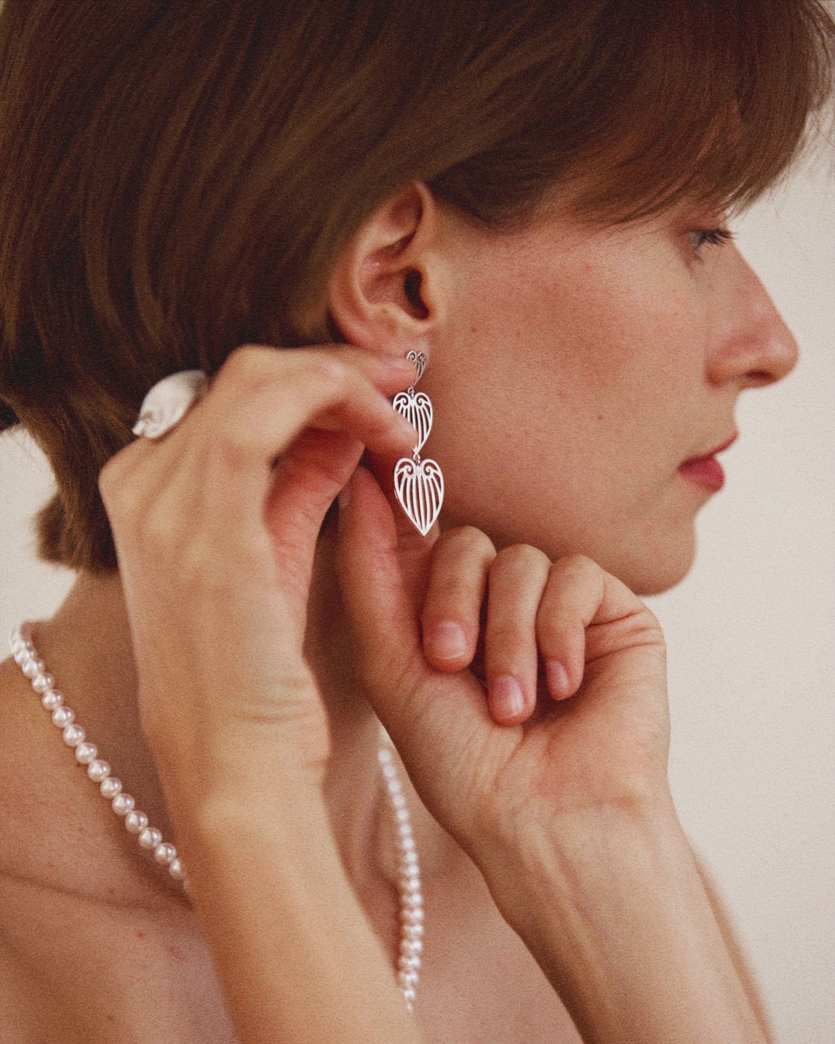Woman wearing heart-shaped earrings and a pearl necklace against a neutral background