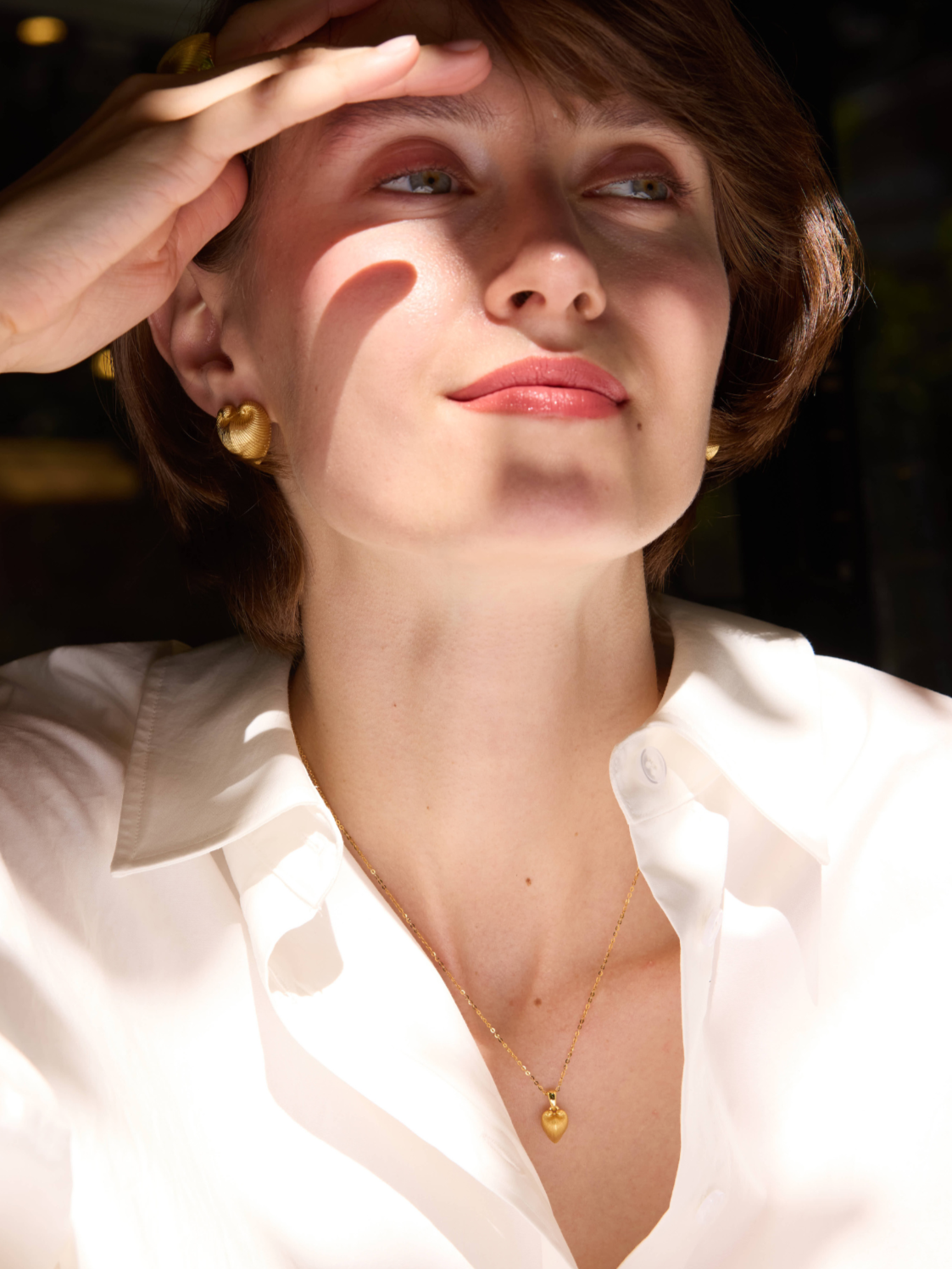 Woman shielding her eyes from the sun with a hand, wearing gold jewelry, with 'Celestes' branding.