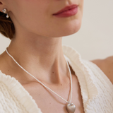 Close-up of a person wearing a pearl necklace with a heart pendant, with 'Celestes' branding.