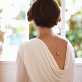 Person wearing a white top with a draped design, standing by a window with a blurred outdoor background.