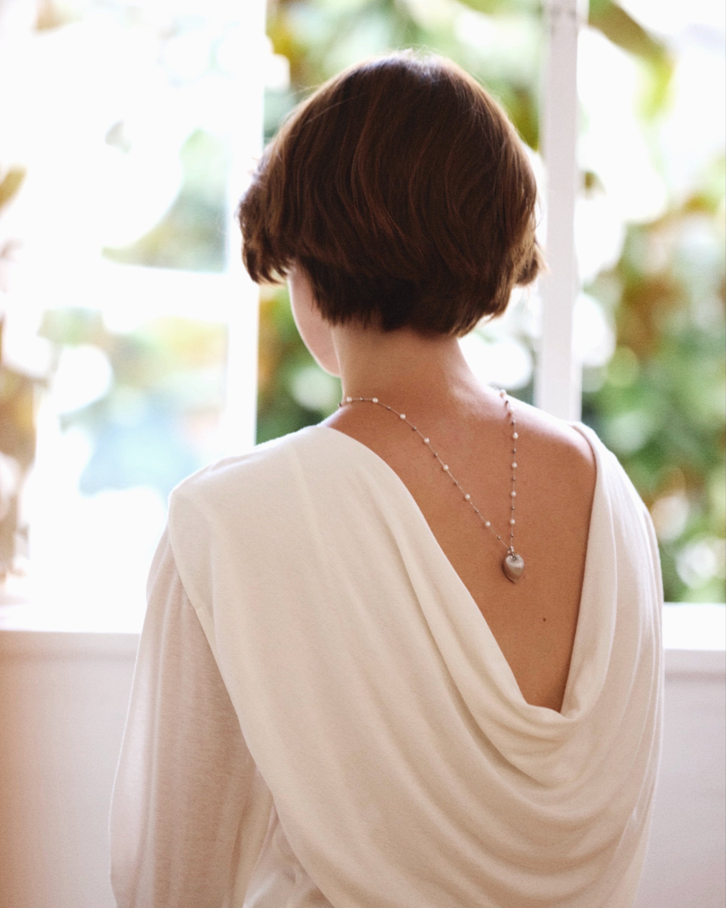 Person wearing a white top with a draped design, standing by a window with a blurred outdoor background.