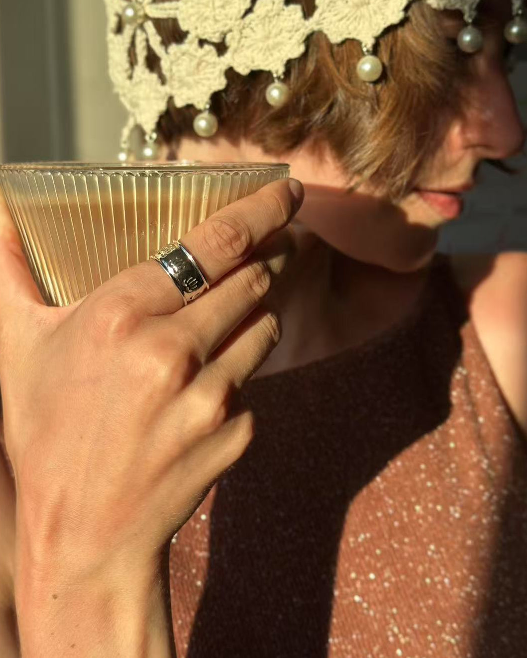 Person holding a glass of coffee with a decorative headpiece and pearl hair accessories.